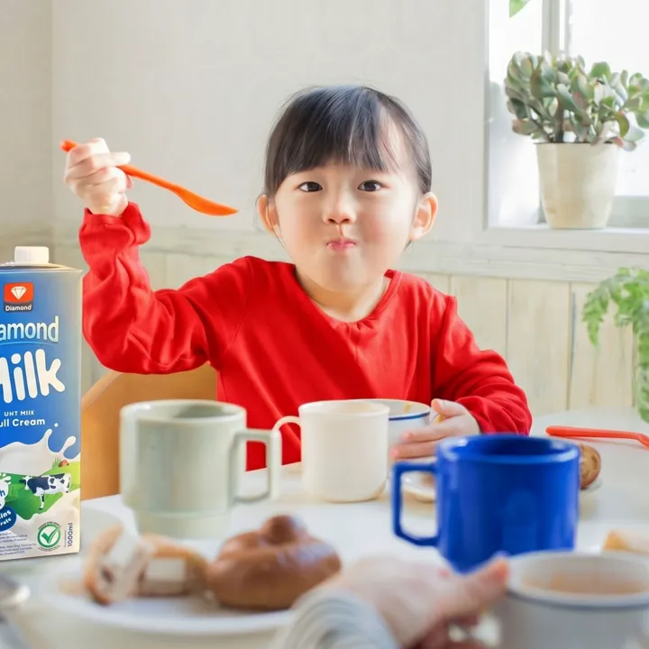 A child at a breakfast table raising a spoon beside a carton of Diamond Milk